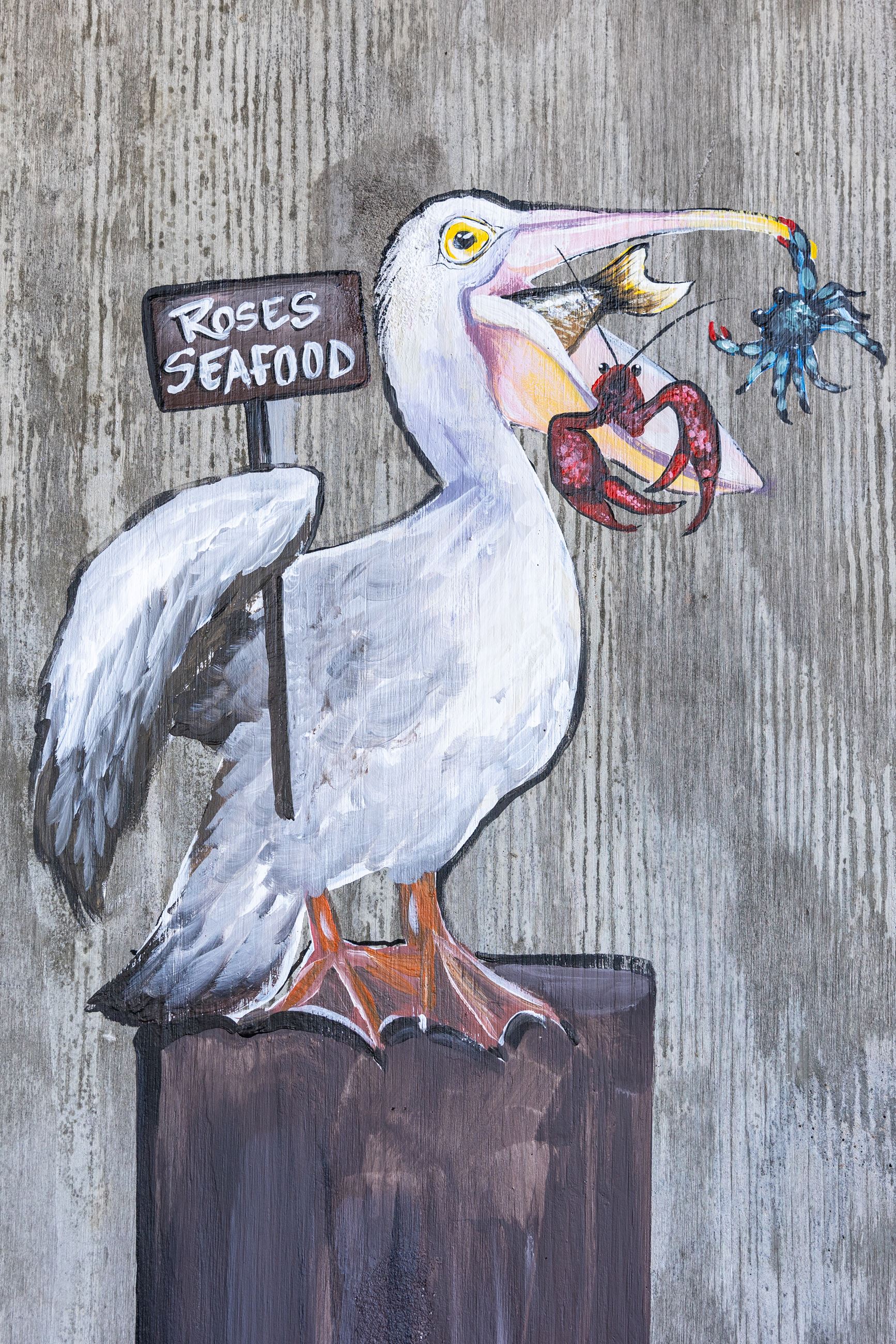 Painted pelican with fish in its mouth holding a sign that says Rose's Seafood